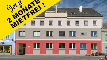 Mehrstöckiges Wohngebäude mit farbiger Fassade, Satteldach und kleinem Balkon im obersten Stockwerk.