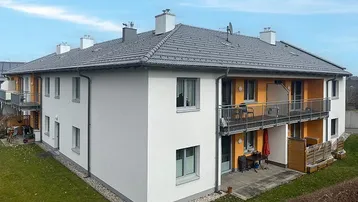 Modernes Mehrfamilienhaus mit weißer Fassade, grauen Dächern und orangefarbenen Akzenten, inklusive Terrasse und Balkonen.