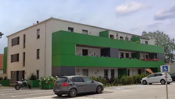Modernes Mehrfamilienhaus mit grüner Fassadengestaltung und barrierefreien Parkplätzen.
