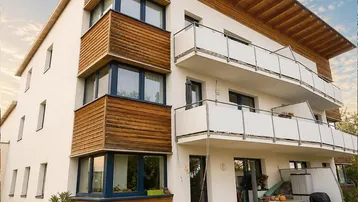 Modernes Mehrfamilienhaus mit weißer Fassade, Holzverkleidung und mehreren Balkonen unter blauem Himmel.