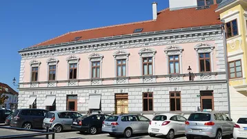 Historisches Gebäude mit rosa Fassade, vielen Fenstern und rotem Ziegeldach, umgeben von parkenden Autos.