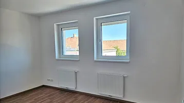 Leeres Zimmer mit zwei Fenstern, Laminatboden und Blick auf die Dächer der Nachbarschaft.