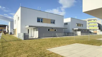 Moderne Reihenhäuser mit weißer Fassade, grünem Rasen und gepflasterter Terrasse an einem sonnigen Tag.
