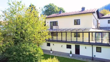 Großzügiges Haus mit verglaster Terrasse und gepflegtem Gartenbereich unter blauem Himmel.