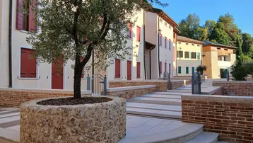 Mediterraner Innenhof mit Olivenbaum, Steinmauern und traditionellen Gebäuden mit roten Fensterläden.