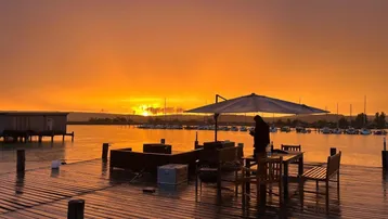 Holzsteg mit Sitzgelegenheiten und Sonnenschirm bei einem wunderschönen Sonnenuntergang über dem See mit Blick auf den Hafen.