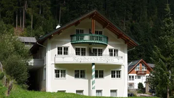 Traditionelles Gebäude mit Balkon und Bergblick, umgeben von grünen Bäumen und Natur.