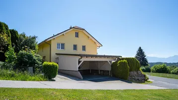 Gelbes Einfamilienhaus mit Carport und Garage in ländlicher Umgebung unter blauem Himmel.