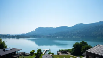 Panoramablick auf den See mit umliegenden Bergen und einigen Häusern am Ufer.