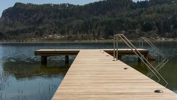 Ein langer Holzsteg führt in den See, umgeben von bewaldeten Bergen unter blauem Himmel.