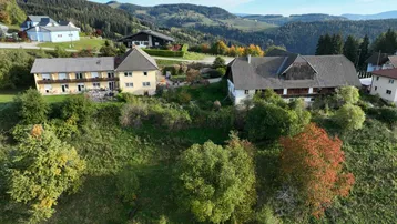 Luftaufnahme einer ländlichen Siedlung mit mehreren Gebäuden und bewaldeten Hügeln im Hintergrund unter blauem Himmel.