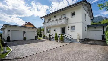 Weißes Einfamilienhaus mit Balkon, Doppelgarage und großem Kiesweg, umgeben von grüner Landschaft.