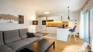 Heller offener Wohnbereich mit moderner Küche, Parkettboden und Zugang zum Balkon.