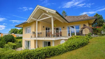 Gelbes Einfamilienhaus mit großem Balkon, umgeben von grüner Hecke und Garten.