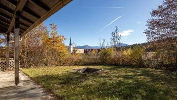 Gartenbereich mit überdachter Terrasse, herbstlicher Vegetation und weitem Blick auf Kirche und Berge.