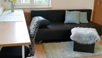Gemütlicher Wohnbereich mit schwarzem Sofa, Teppich und einem kleinen Esstisch am Fenster.