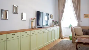 Helles Wohnzimmer mit langer, hellgrüner Schrankwand, Fernseher und großen Fenstern.