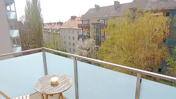 Kleiner Balkon mit Holztisch und Blick auf die umliegenden Gebäude und Bäume.