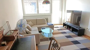 Gemütliches Wohnzimmer mit beigem Ecksofa, Fernseher und gemustertem Teppichboden.