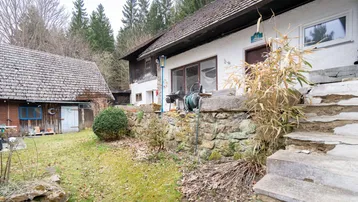 Rustikales Bauernhaus mit Steinterrasse und Gartenbereich, umgeben von Bäumen und Natur.