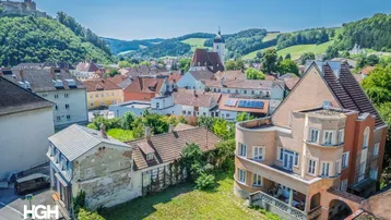 Historisches Gebäude in einer malerischen Stadtansicht mit Burg und grünen Hügeln im Hintergrund.