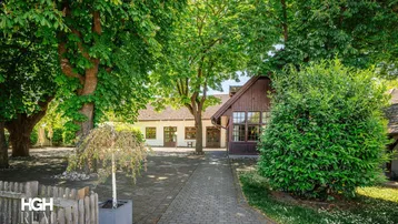 Ein charmantes Gebäude mit traditioneller Architektur, umgeben von üppigem Grün und hohen Bäumen, mit einem gepflasterten Weg zum Eingang.