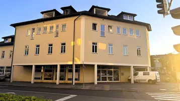 Helles Eckgebäude mit moderner Architektur und Geschäften im Erdgeschoss, beleuchtet von der Abendsonne.