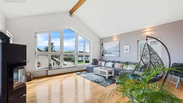 Helles Wohnzimmer mit hohen Decken, Holzbalken, Kaminofen und großen Fenstern mit Weitblick.