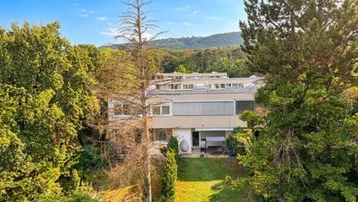 Gebäudeansicht von hinten mit Gartenbereich und umgebenden Bäumen, im Hintergrund Hügel.