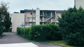 Mehrfamilienhaus mit mehreren Etagen, Außentreppen und begrüntem Vorgartenbereich.