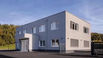 Modernes Bürogebäude mit grauer Fassade und großen Fenstern, umgeben von Natur unter blauem Himmel.