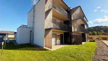 Modernes Mehrfamilienhaus mit Balkonen, Holzlamellenfassade und Grünflächen unter blauem Himmel.