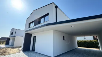 Modernes Einfamilienhaus mit weißer Fassade, schwarzem Dach und integriertem Carport unter blauem Himmel.