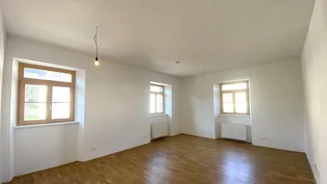 Helles Zimmer mit Parkettboden, weißen Wänden und mehreren Fenstern, bereit zur individuellen Gestaltung.