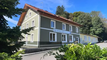 Traditionelles Gebäude mit grüner Fassade und rotem Ziegeldach, umgeben von Bäumen und einem Parkplatz.