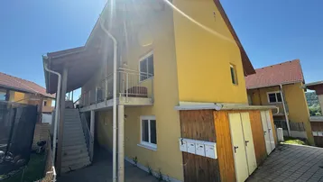 Gelbes Mehrfamilienhaus mit Balkonen, Außentreppe und Schuppen unter strahlendem Sonnenschein.