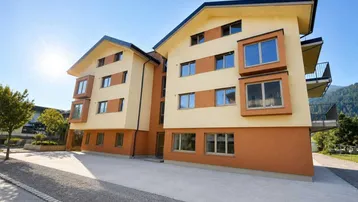 Modernes Mehrfamilienhaus mit gelb-orangener Fassade und großen Fenstern in sonniger Lage mit Bergblick.