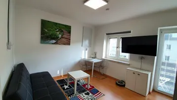 Helles Wohnzimmer mit Sofa, Fernseher, Schreibtisch und Zugang zu einem Balkon.