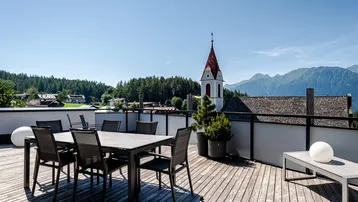 Sonniger Terrassenbereich mit Esstisch und Lounge-Möbeln, bietet einen weiten Blick auf die Berge und das Dorf.