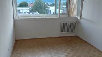 Heller Raum mit Parkettboden, großem Fenster und Blick auf die Berge und die Umgebung.