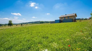 Ein im Bau befindliches Haus in einer weiten grünen Landschaft unter blauem Himmel mit blühenden Wiesen.