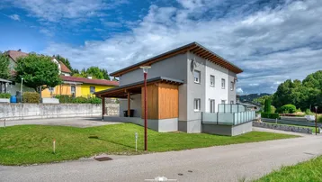 Modernes Haus mit grauer Fassade, Holzverkleidung und Carport in ländlicher Umgebung.