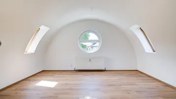 Heller Raum im Dachgeschoss mit gewölbter Decke, Rundfenster und Holzboden.