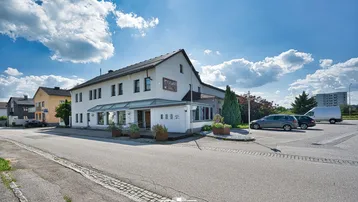 Weißes Gebäude mit Satteldach und großem Parkplatz unter blauem Himmel.