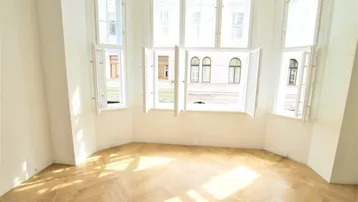 Helles Zimmer mit Fischgrätparkett und einem großen Erkerfenster, das viel Tageslicht hereinlässt.