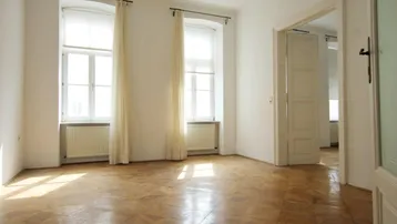 Helles Zimmer mit Fischgrätparkettboden und hohen Fenstern, das viel Tageslicht hereinlässt.