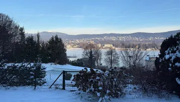 Winterliche Landschaft mit schneebedecktem Garten, See und Bergen im Hintergrund unter blauem Himmel.