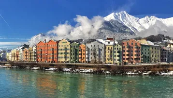 Panoramablick auf eine historische Stadt am Flussufer mit bunten Häusern und schneebedeckten Bergen im Hintergrund.