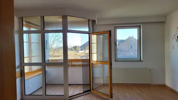Helles Zimmer mit Laminatboden und Zugang zu einem verglasten Balkon, bietet viel Tageslicht.