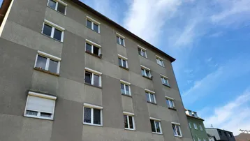 Die Fassade des Mehrfamilienhauses mit mehreren Fenstern und Rollläden unter blauem Himmel.